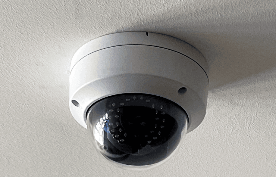 Ceiling-mounted dome security camera with a white housing and dark tinted lens, installed indoors and angled downward for surveillance.
